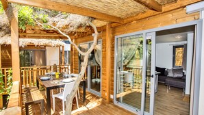 Eine Holzterrasse mit Esstisch und Stühlen, ein Reetdach und große Schiebegläser zu einem Wohnbereich.