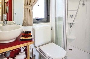 A bathroom with a white sink, toilet, and shower. There is a window with curtains, and a shelf with toiletries.