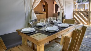 Ein Holztisch ist gedeckt mit Tellern, Gläsern und Besteck, umgeben von Holzstühlen, in einem Zeltbereich für Mahlzeiten im Freien.