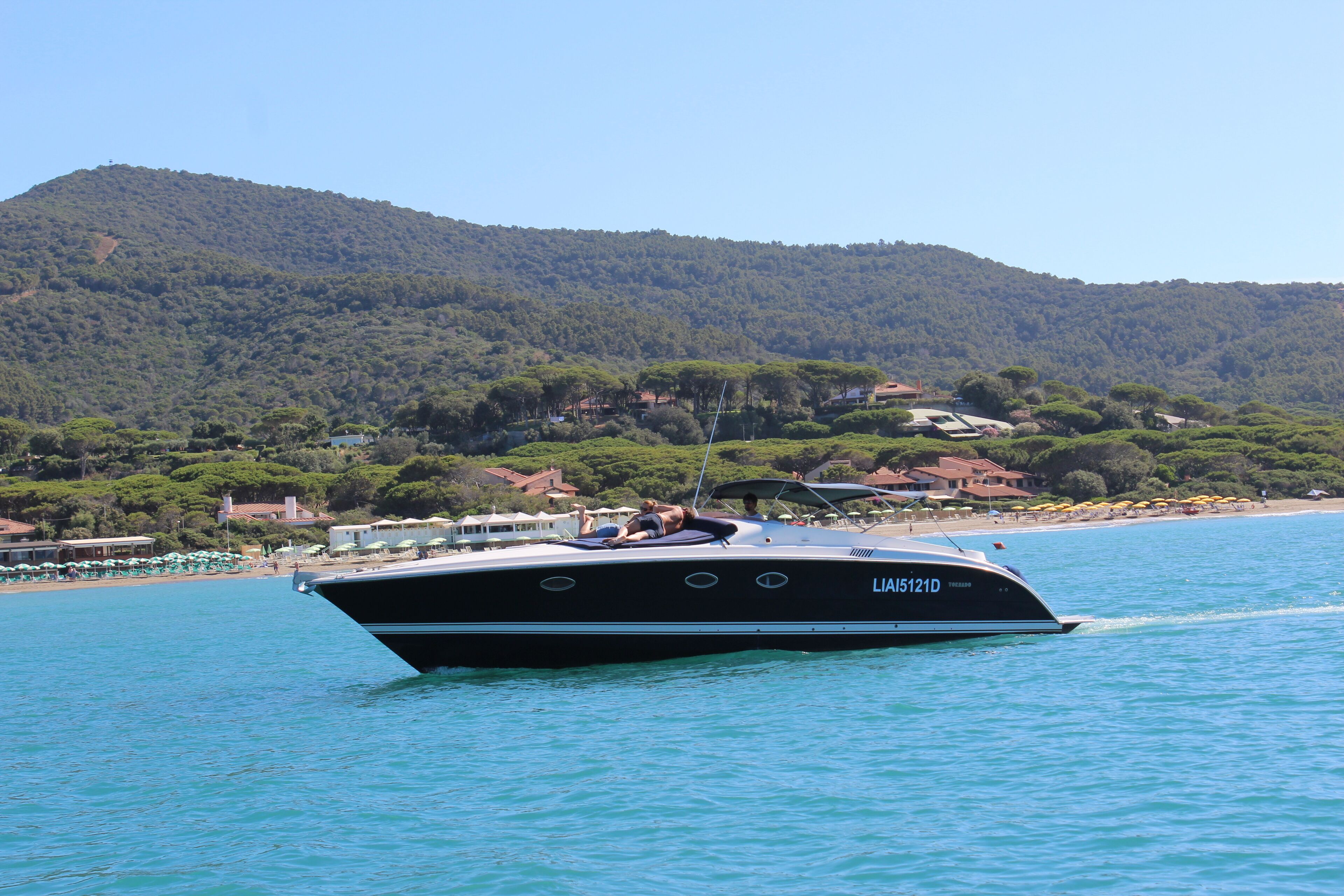 Ein Boot auf ruhigem Wasser, mit Bergen im Hintergrund und einem Strand mit Sonnenschirmen.