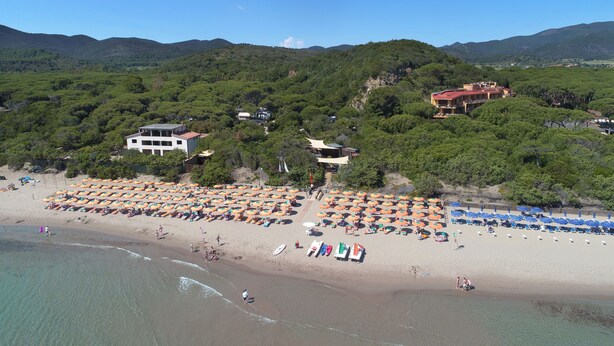 Ein Strand mit Reihen von Sonnenschirmen und Liegestühlen, ein Gebäude mit rotem Dach und eine gebirgige Kulisse.