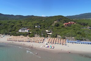 Une plage bordée de rangées de parasols et de chaises longues, un bâtiment avec un toit rouge et, en arrière-plan, un paysage montagneux.