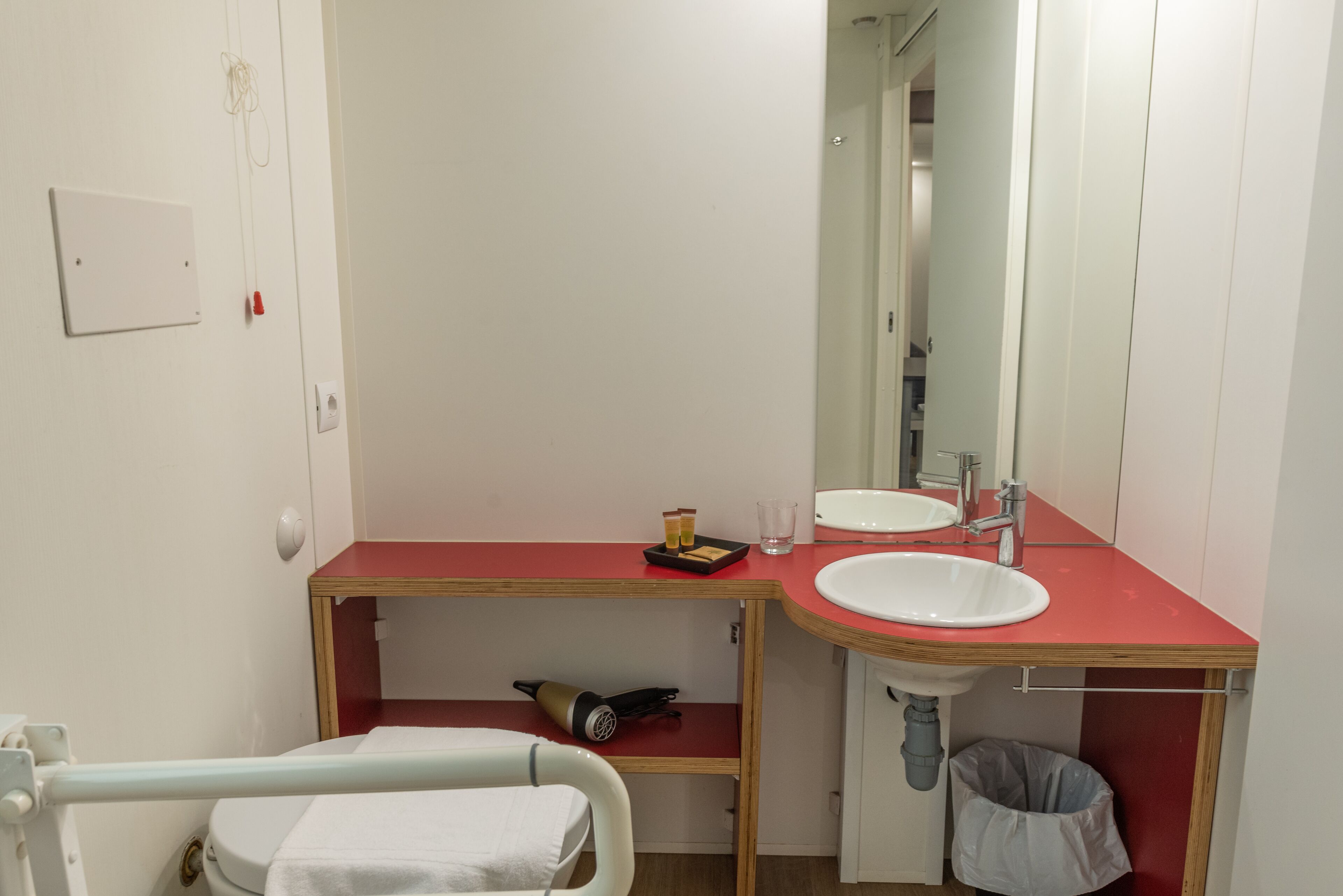 Un baño con encimera roja, lavabo blanco y espejo. Hay un perchero, un cubo de basura y un secador de pelo en la encimera.