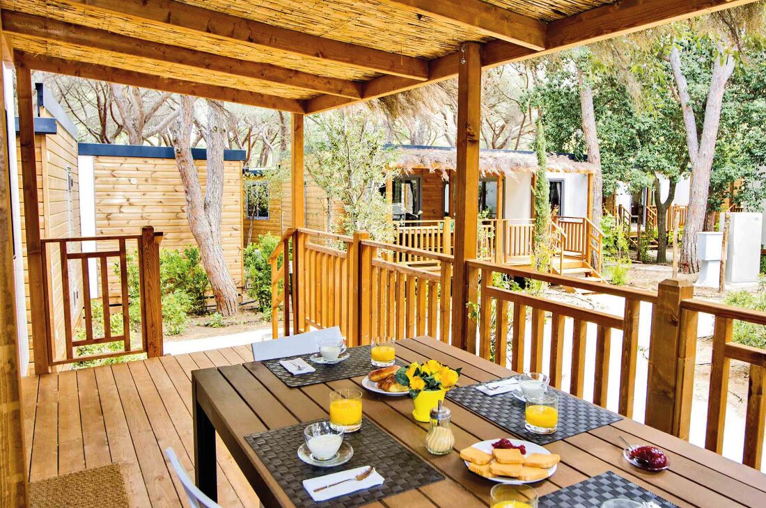 Una terraza de madera con el desayuno servido, incluyendo platos, vasos y un bol de frutas.
