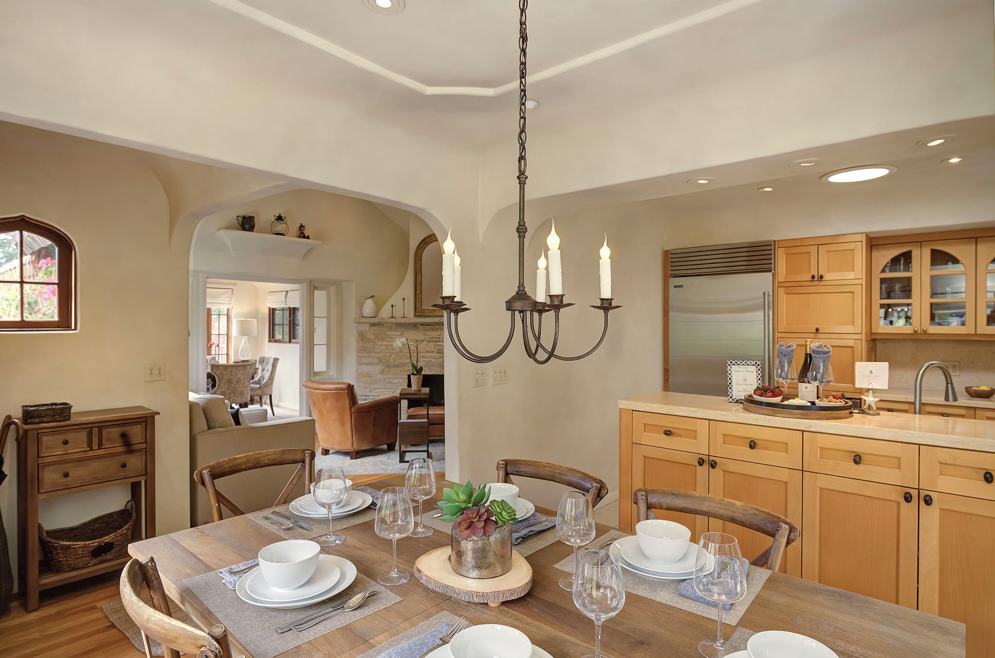 Un comedor con una mesa de madera dispuesta para seis personas, rodeada de sillas de madera. Un candelabro cuelga sobre la mesa, y se distingue una cocina al fondo.
