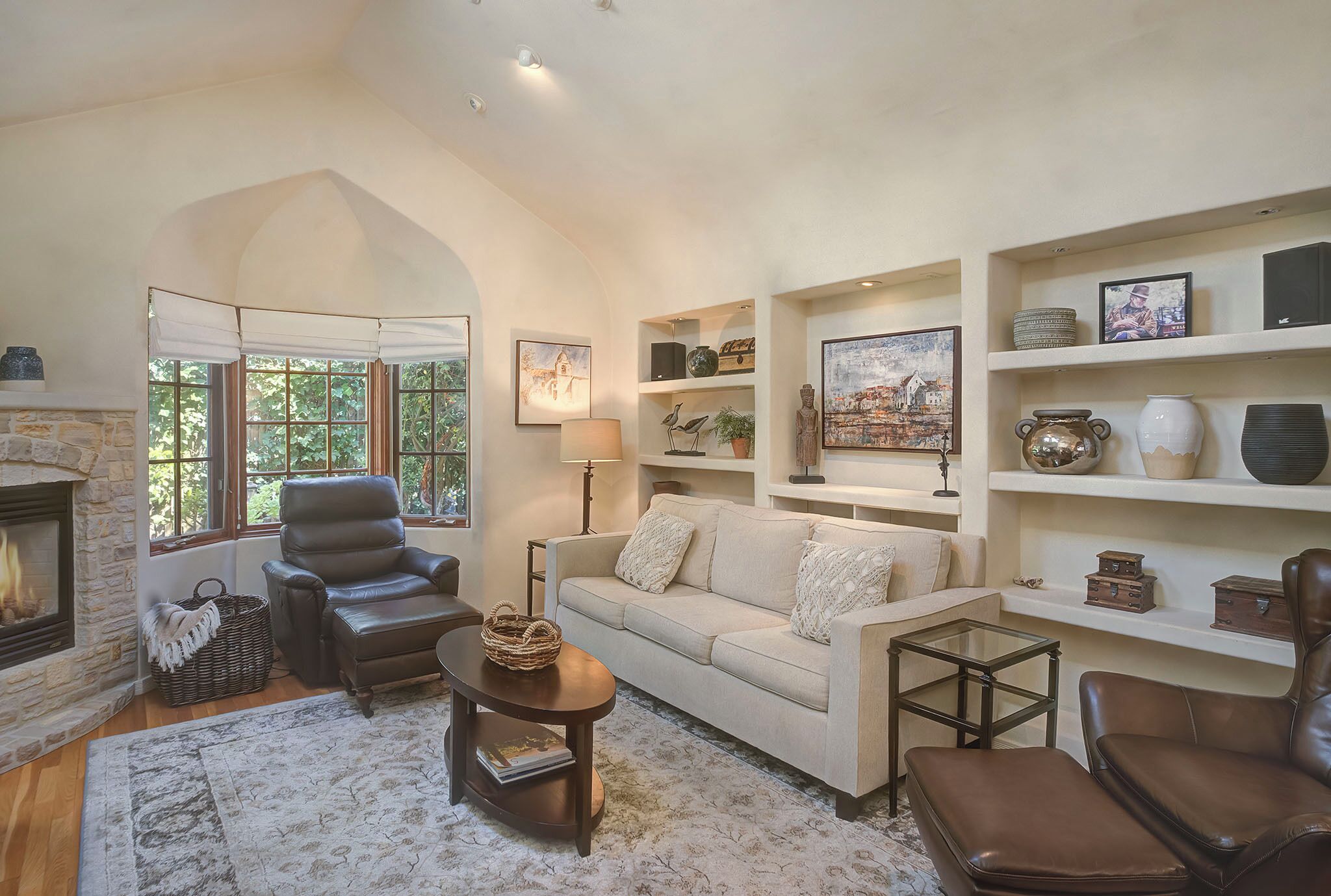 A cozy living room with a fireplace, leather armchairs, a sofa, and a coffee table. There are shelves with decorative items and a large window with a view of greenery.