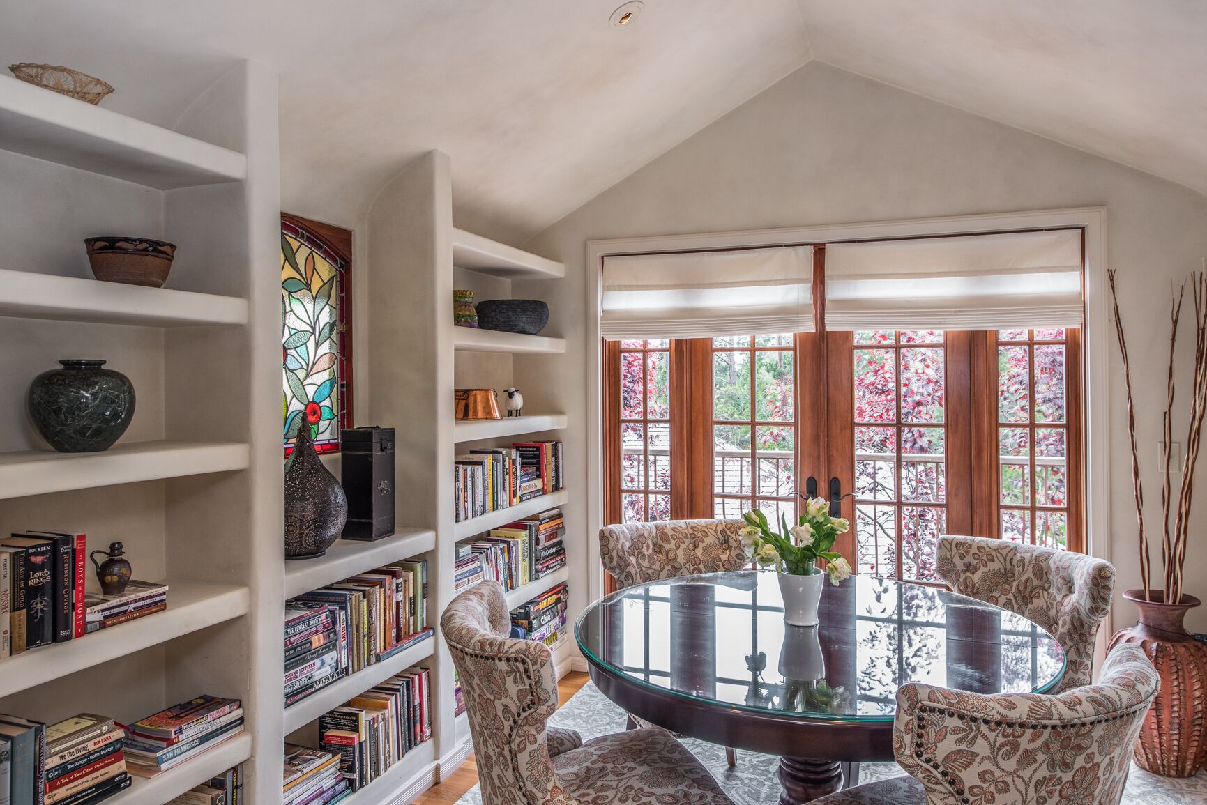 A room with a round glass table surrounded by patterned chairs, a bookshelf filled with books, and a stained glass window.