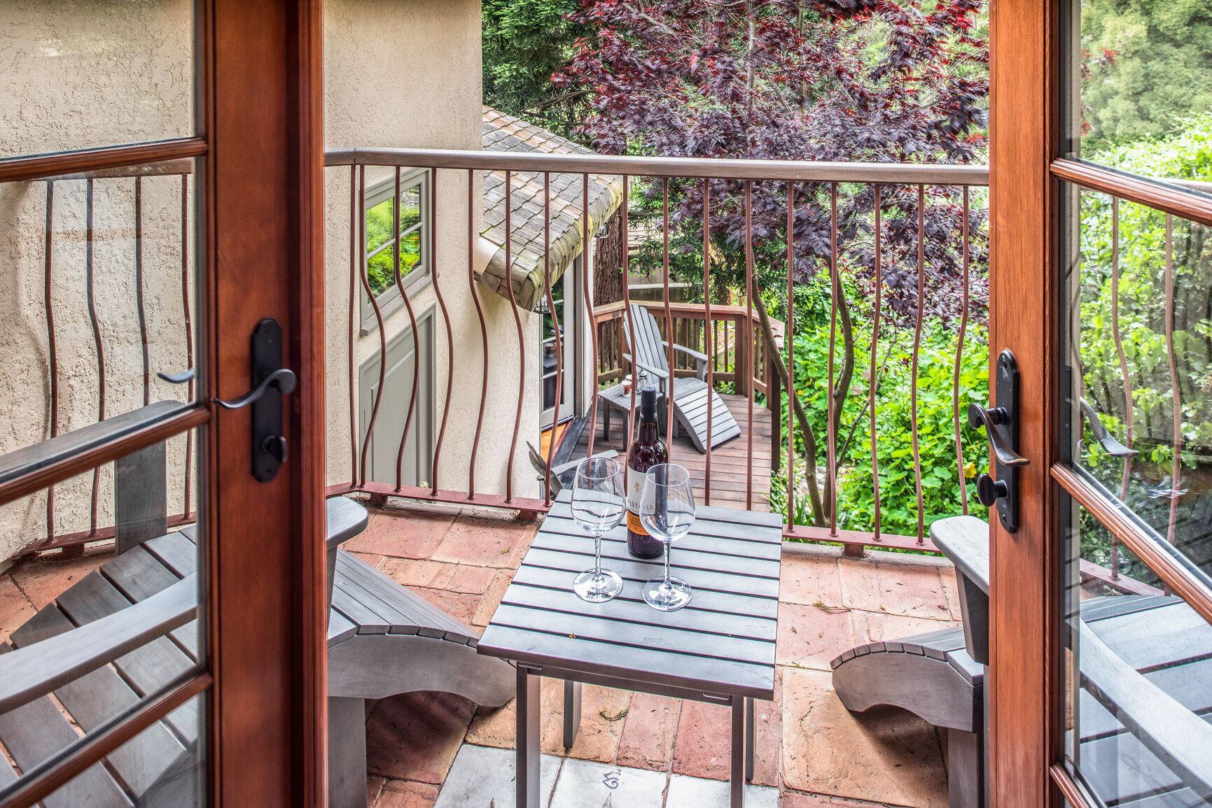 Une terrasse extérieure avec une table et des chaises, une bouteille de vin et une vue sur un jardin arboré avec une terrasse en bois.