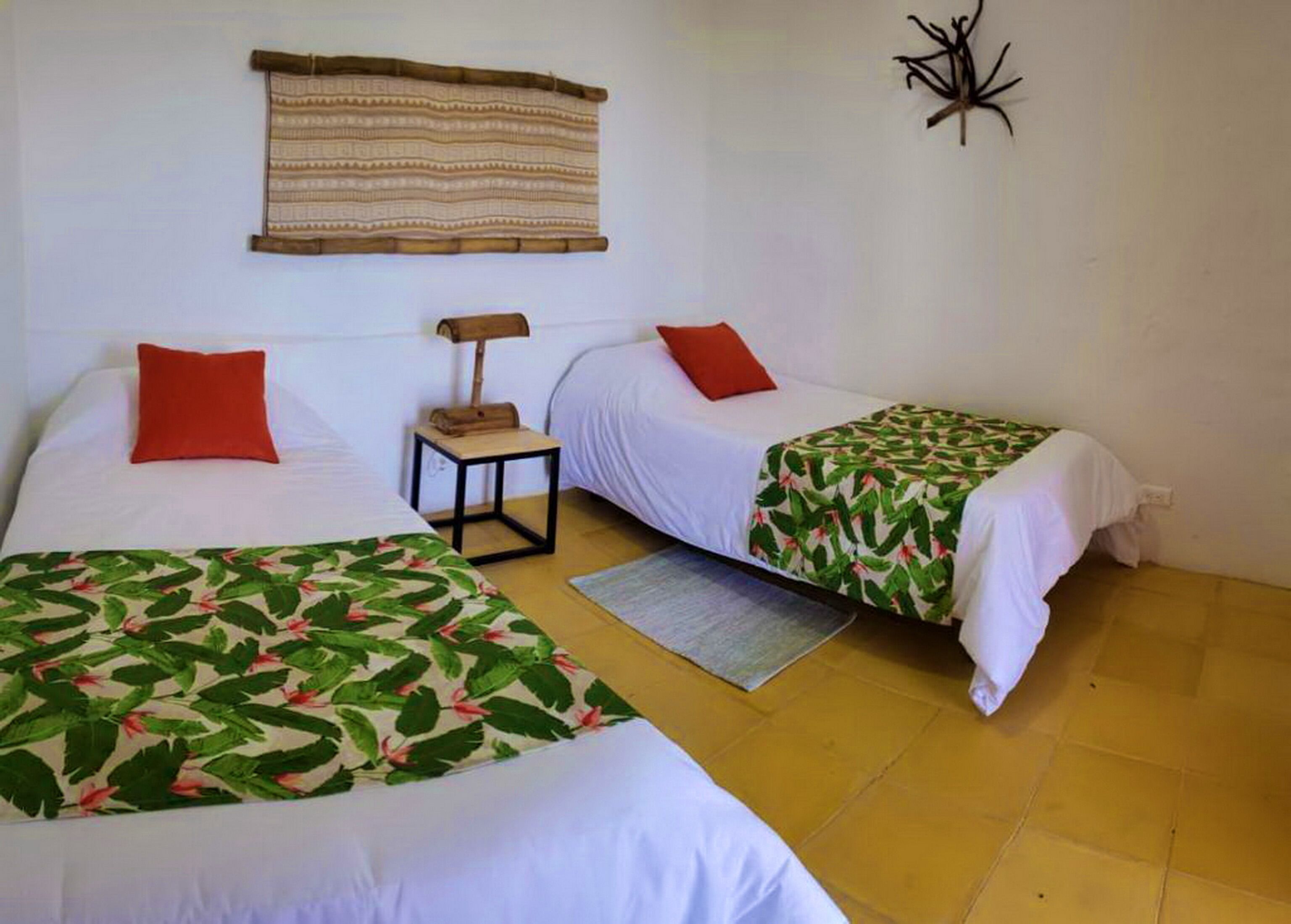 Quarto com duas camas, cada uma com travesseiro vermelho e colcha florida. Há uma pequena mesa com uma luminária e uma tapeçaria tecida na parede.