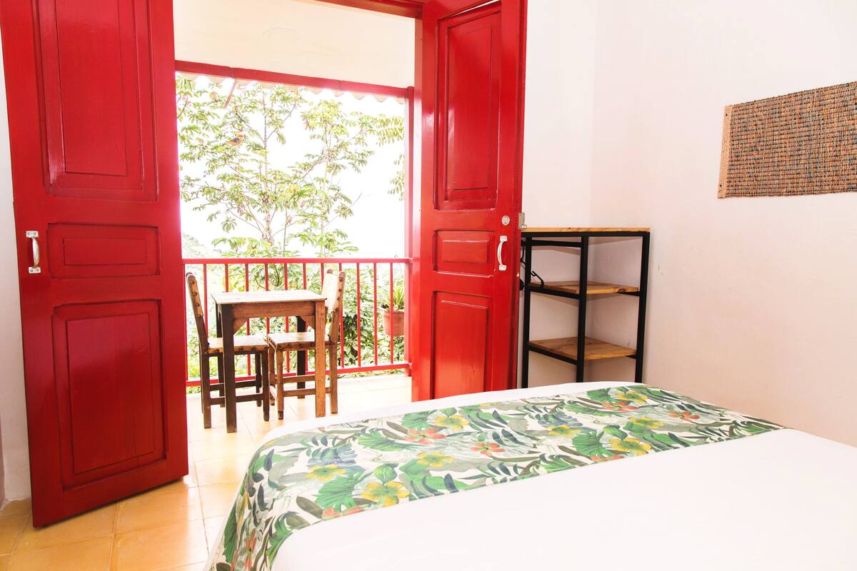 Ein Zimmer mit einem Bett, einem kleinen Tisch und einem Stuhl. Es gibt einen Balkon mit Geländer und Blick auf Bäume.