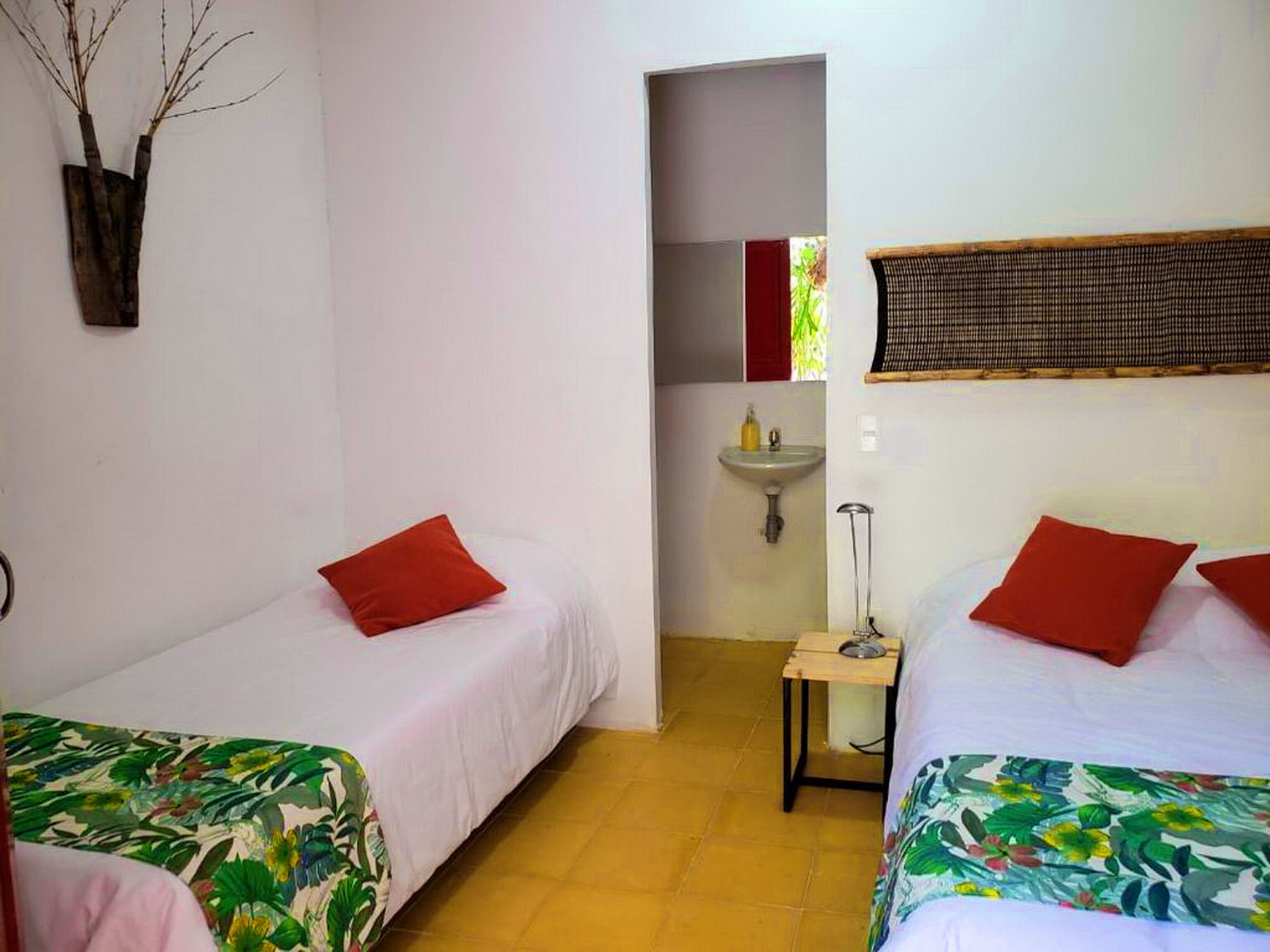 A room with two beds, each with a red pillow and a floral bedspread. There is a small table with a lamp and a bottle of soap. A sink is visible in the background.