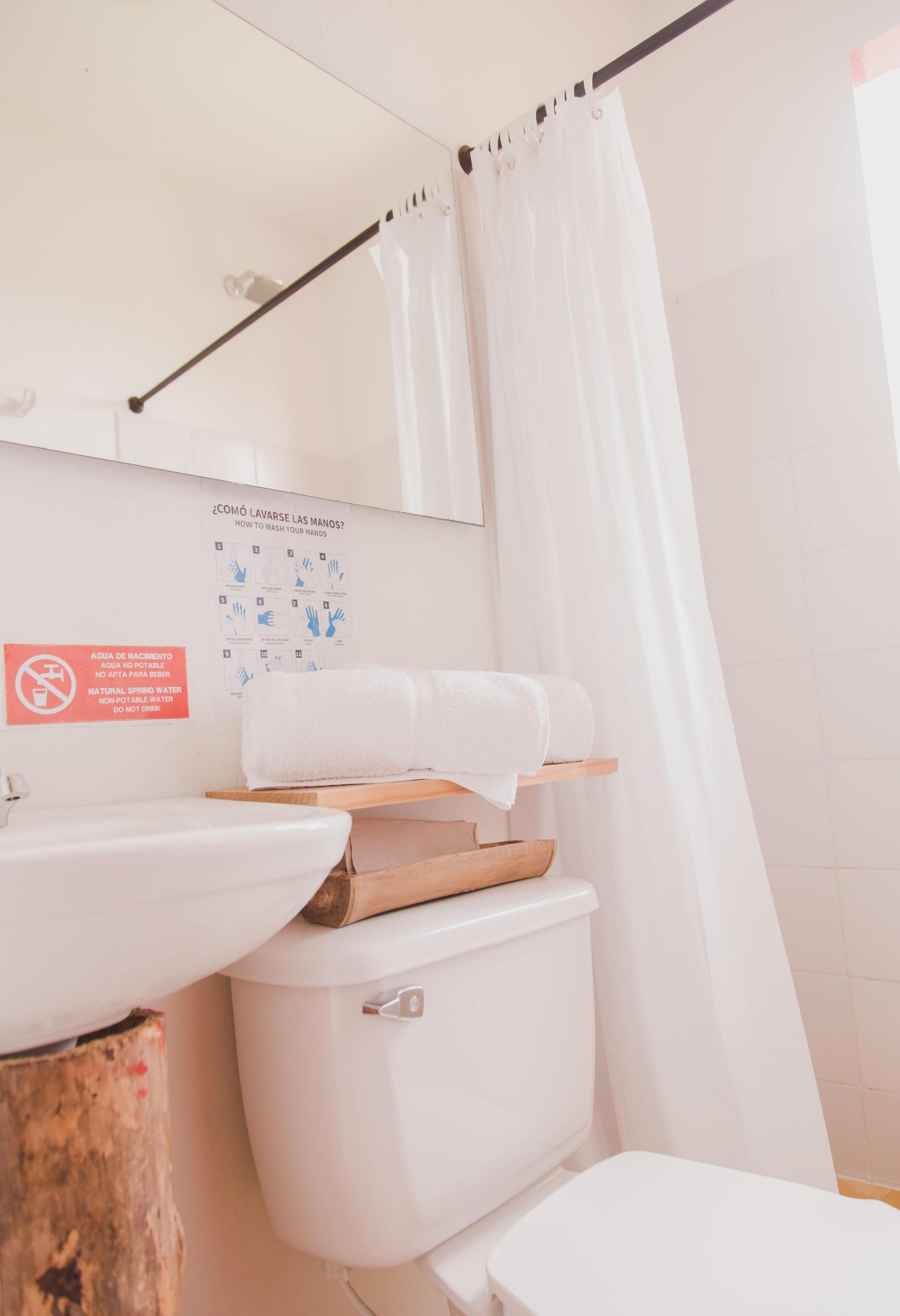 A bathroom with a sink, toilet, and shower curtain. There is a sign with handwashing instructions and a no-drink sign.
