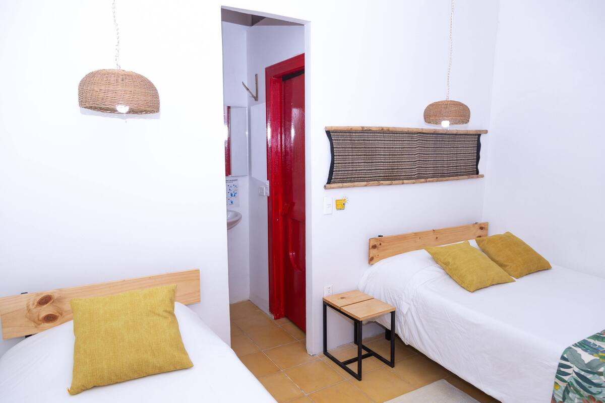 Ein kleines, sauberes Zimmer mit zwei Betten, einem Holz-Kopfteil, einem kleinen Tisch und einer Pendelleuchte.