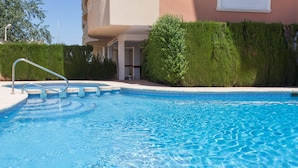 Une piscine avec un escalier en métal, entourée d’une haie et bordée d’un bâtiment doté d’un balcon.