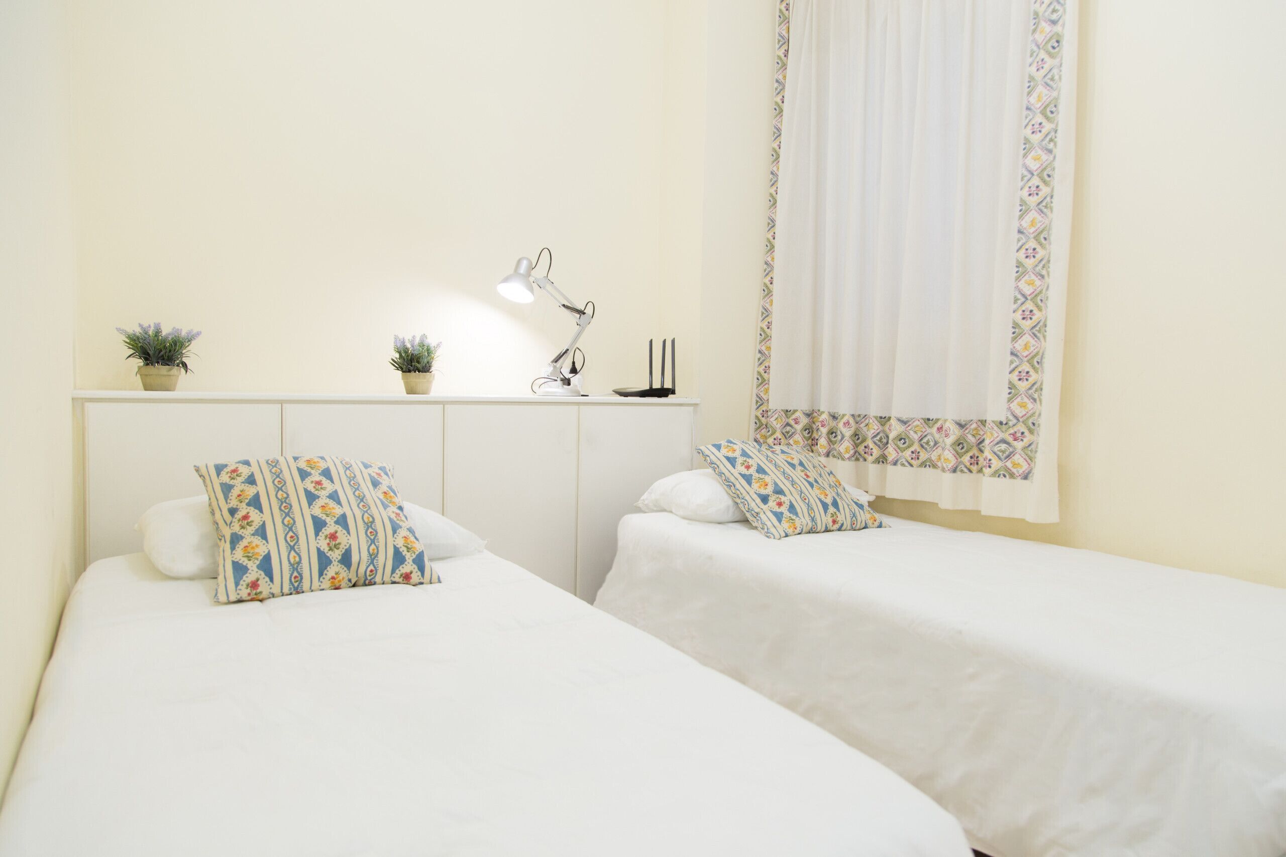 A bedroom with two beds, a desk with a lamp, and two potted plants.