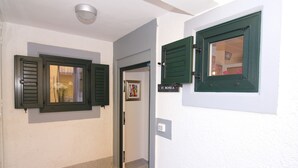 Un couloir avec une porte donnant sur une pièce, une petite fenêtre avec des volets et un luminaire fixé au mur.