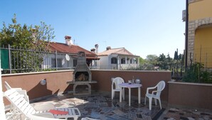Une terrasse extérieure avec un sol carrelé en pierre, des chaises en plastique blanches, une petite table ronde et un barbecue en pierre.