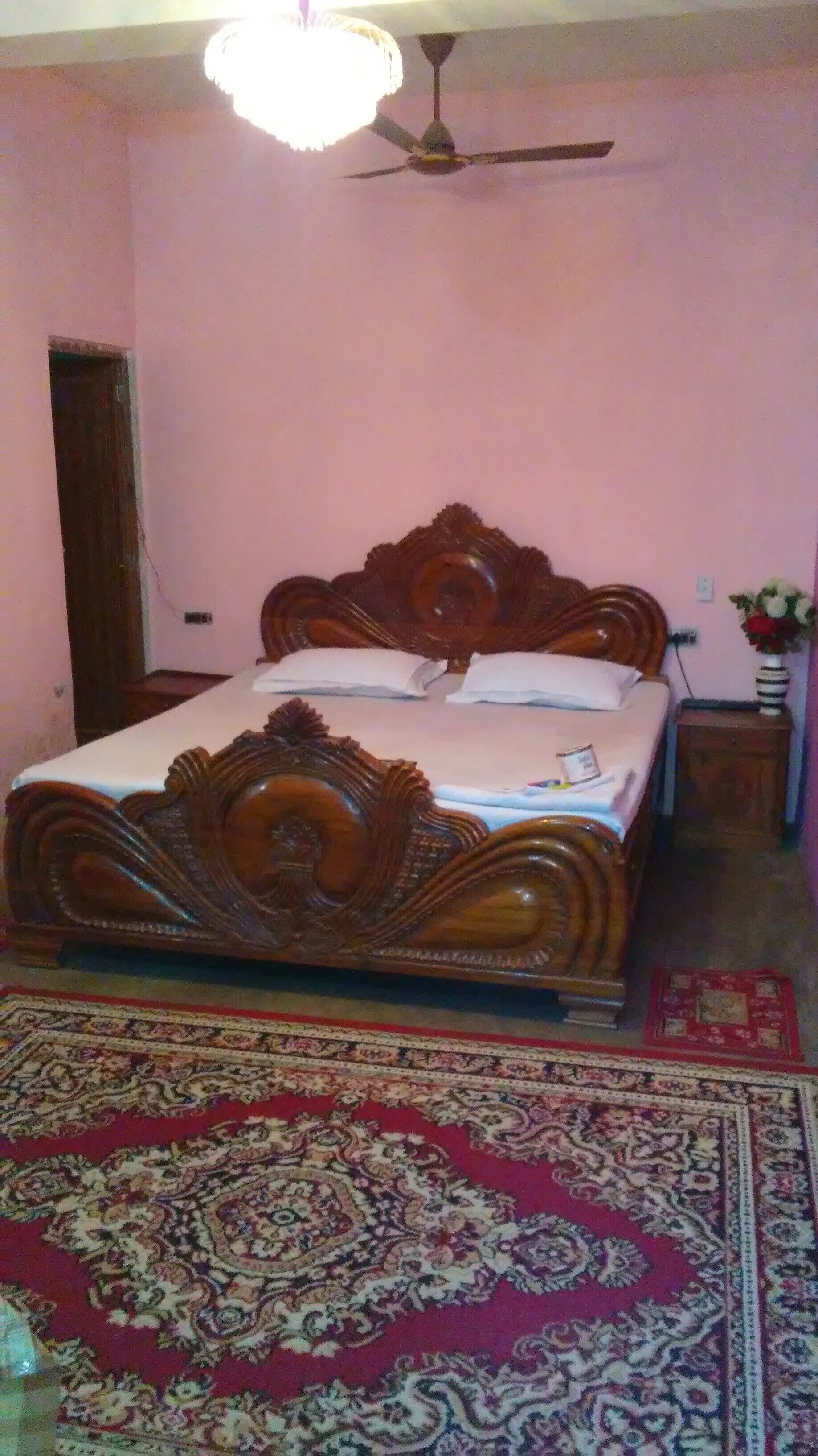 A bedroom with a wooden bed, a ceiling fan, a nightstand with a vase, and a patterned rug.
