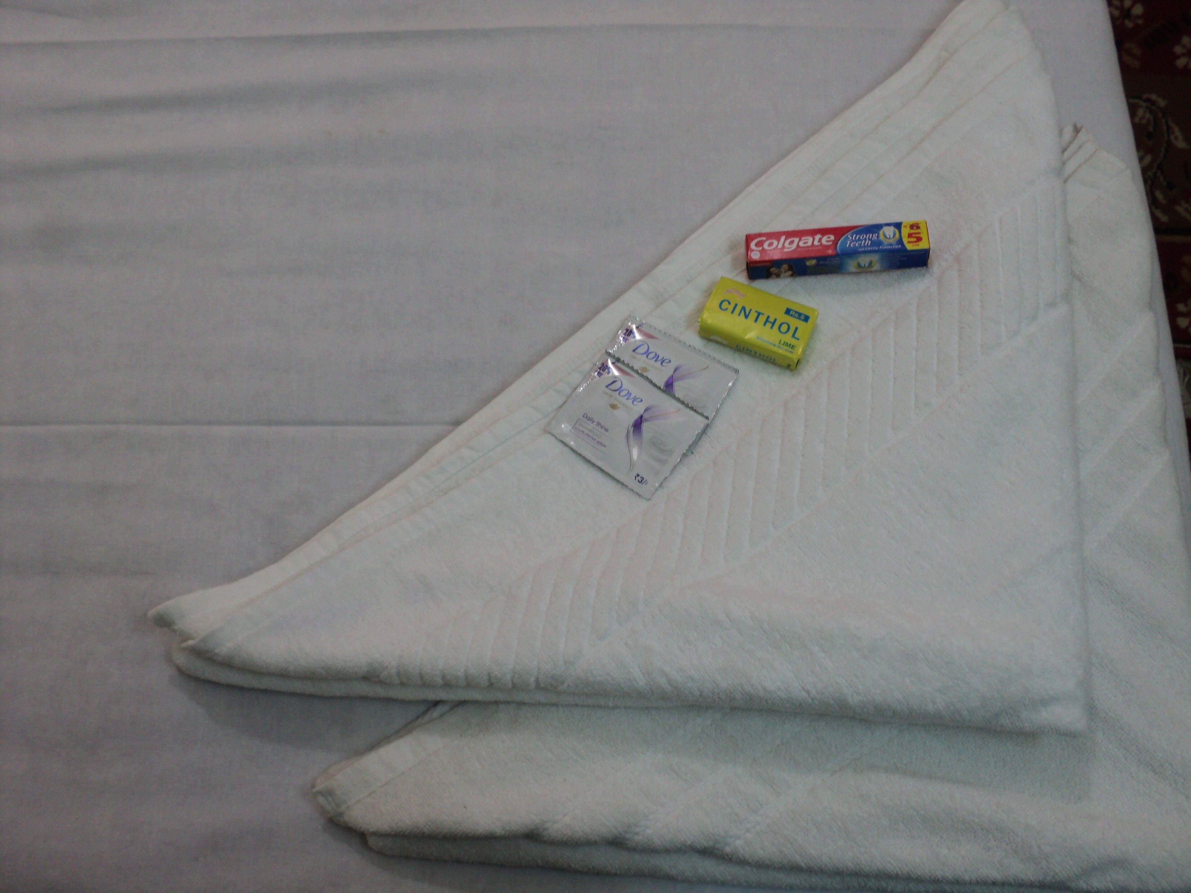 Una cama con una manta blanca, un paquete de toallas higiénicas Dove, un paquete de pasta de dientes Colgate y un paquete de enjuague bucal Cinthol.