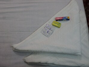 Una cama con una manta blanca, un paquete de toallas higiénicas Dove, un paquete de pasta de dientes Colgate y un paquete de enjuague bucal Cinthol.