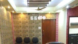 Uma sala de estar com um sofá, uma poltrona, uma mesa de centro e um tapete estampado. Há quatro cadeiras encostadas à parede e um ventilador de teto.