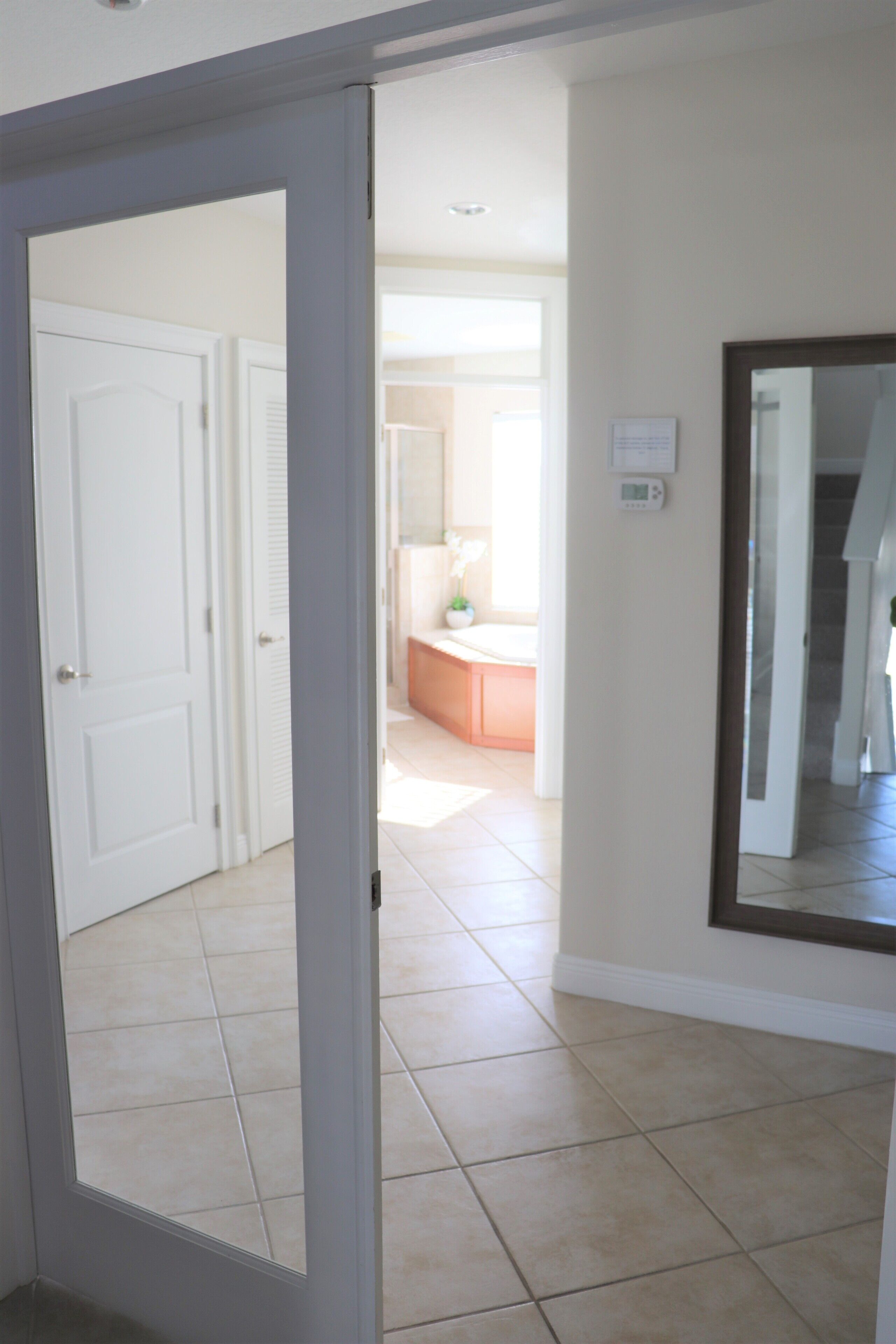 A hallway with a white door, a mirror, and a tiled floor leading to a bathroom with a sink and a toilet.