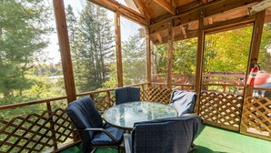A covered outdoor seating area with a glass table and chairs, surrounded by a wooden fence and a view of trees and a lake. - Republic Island -Summer Paradise – Your Private Island Awaits! (Republic)