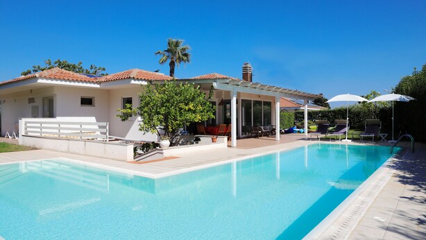 Una piscina con un portico coperto, una palma e un edificio con tetto in tegole.