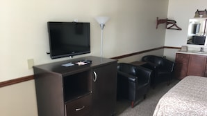 A hotel room with a bed, a desk with a television, a lamp, and two chairs. - Flag's West Motel (Downey)