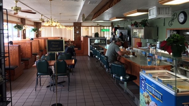 A restaurant with booths and tables, a counter with a menu, and a clock on the wall. - Flag's West Motel (Downey)