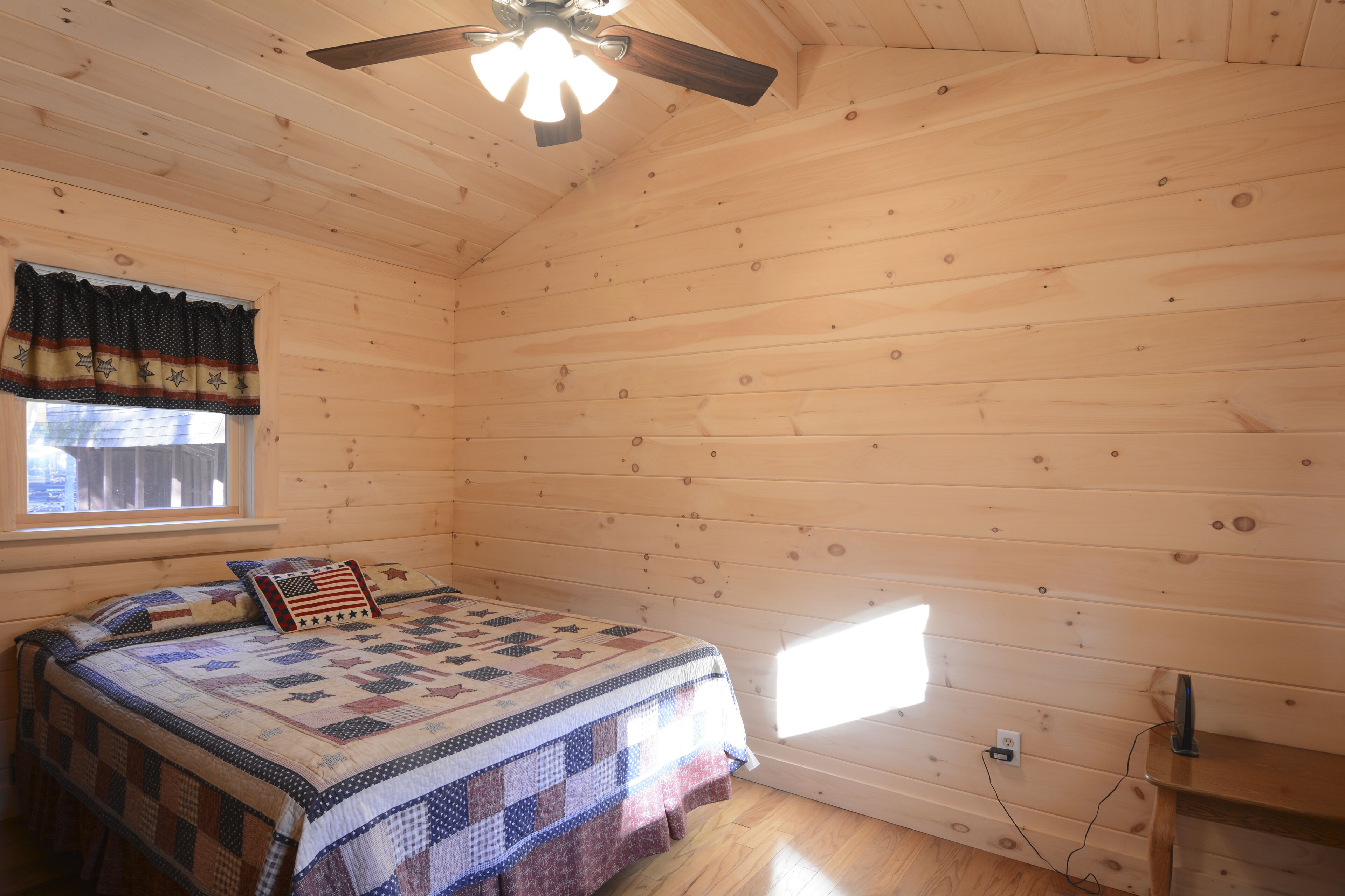 A wooden cabin bedroom with a bed, a ceiling fan, a window with curtains, and a small window on the wall.