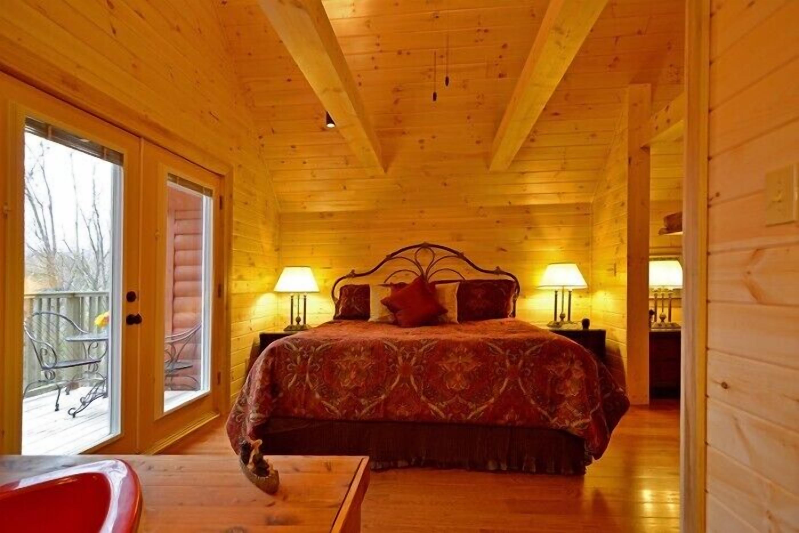 A wooden cabin bedroom with a large bed, two bedside lamps, and a view through a sliding glass door to an outdoor area.