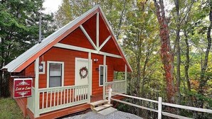 A small, single-story cabin with a porch, surrounded by trees.