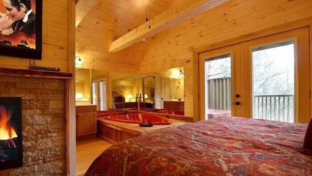 A cozy cabin-style room with a large bed, a fireplace, a stone wall, a wooden ceiling, and a large window.