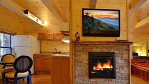 A cozy cabin interior with a stone fireplace, wooden ceiling beams, a kitchen area, and a dining space.