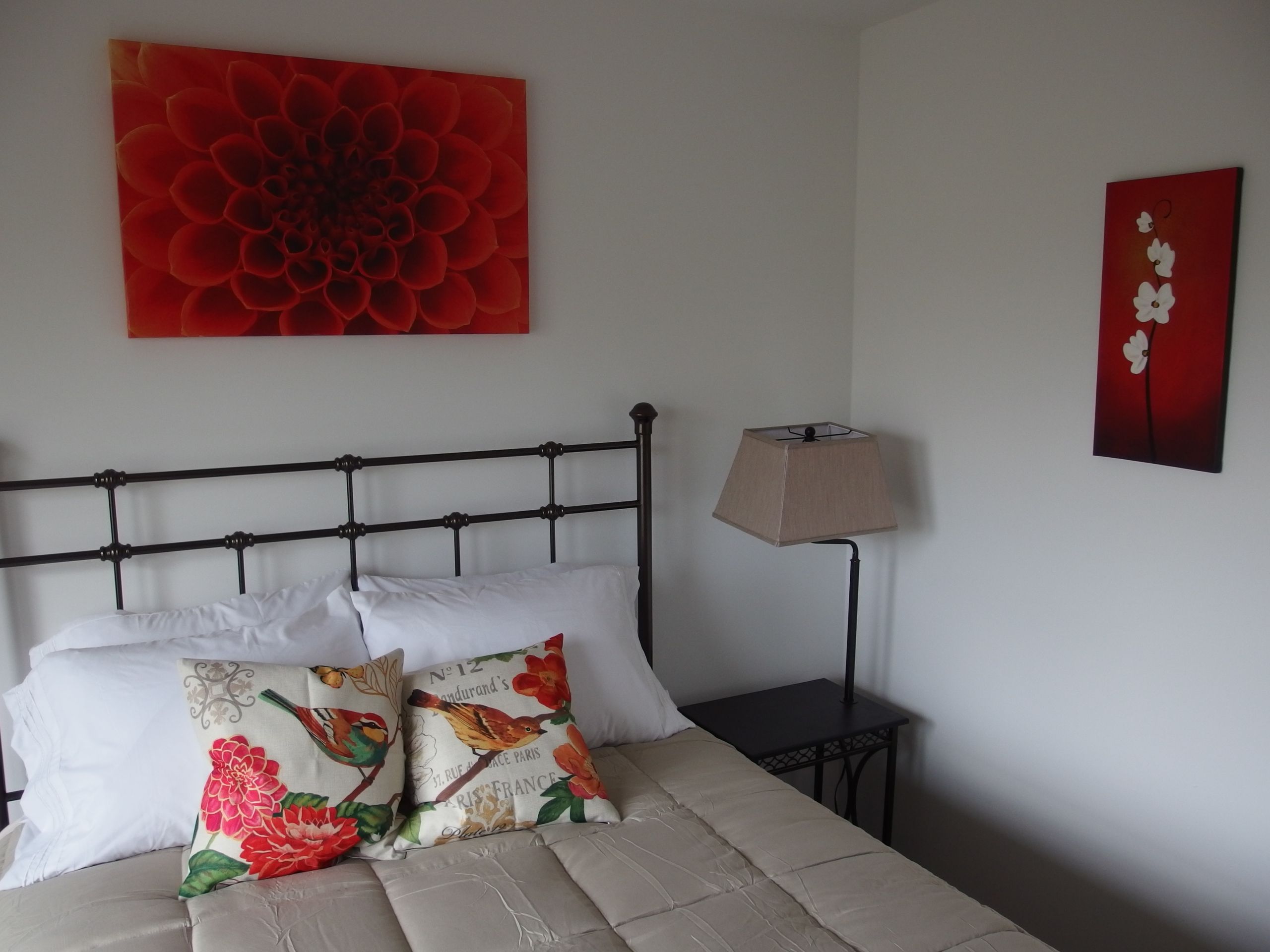 Un lit avec un cadre en métal, des oreillers blancs et une couvre-lit beige. Une lampe est posée sur une petite table à côté du lit.