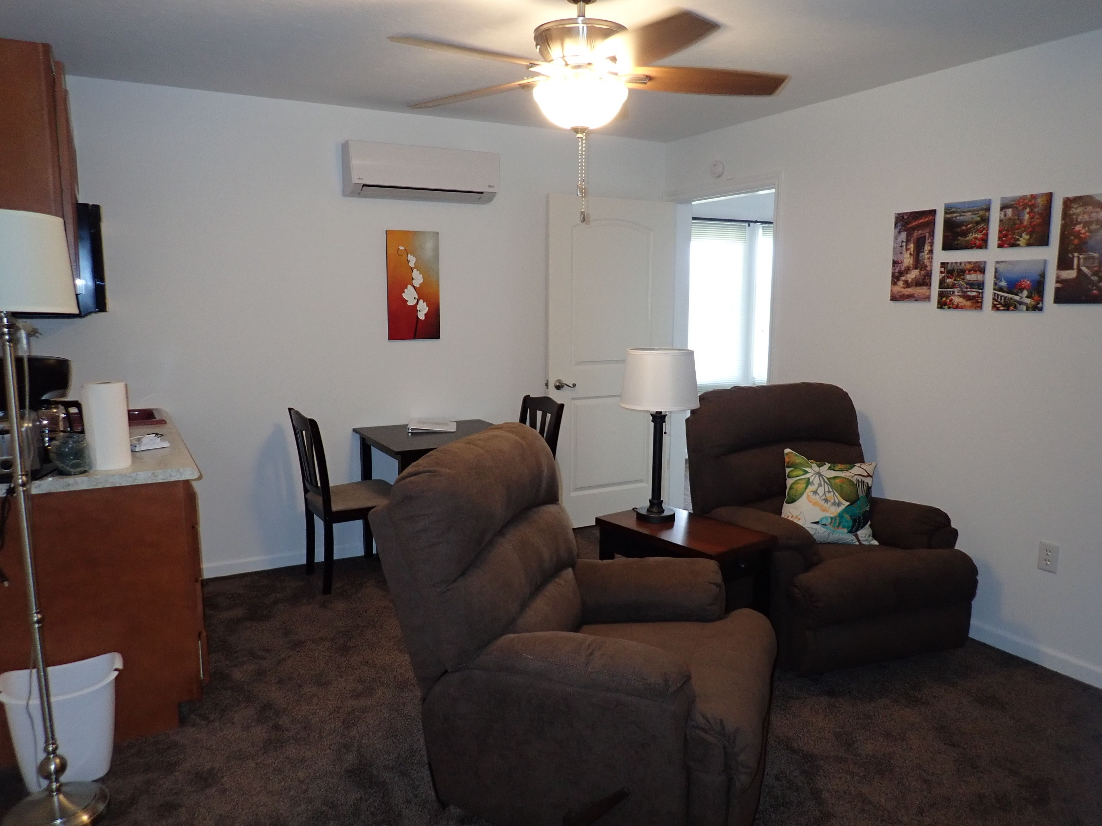 A living room with a ceiling fan, a dining table with chairs, a sofa, a lamp, and a wall-mounted air conditioner.