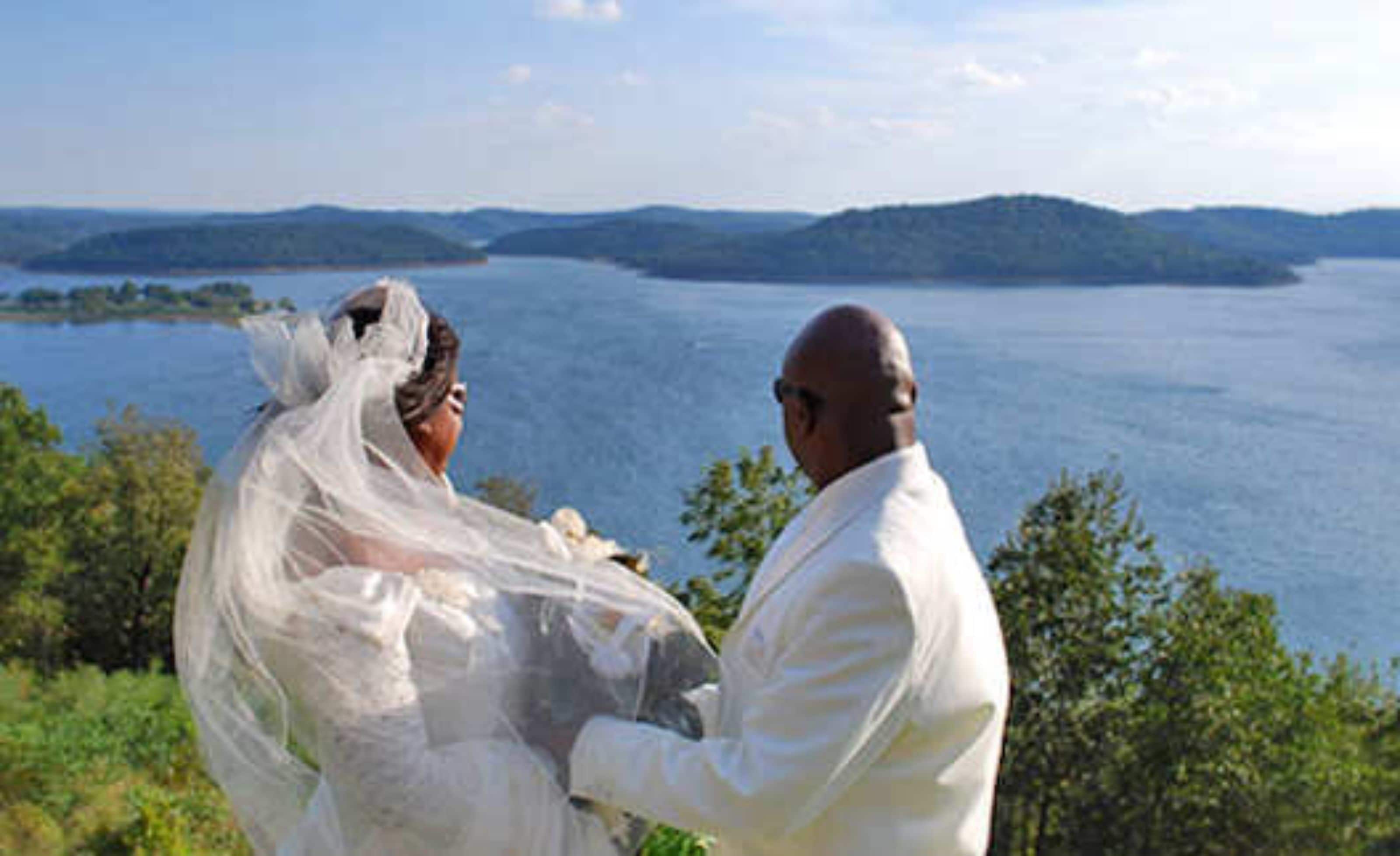 A couple in wedding attire by a lake with a view of distant hills.