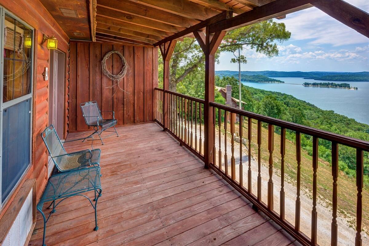 A wooden deck with two metal chairs and a lantern, overlooking a lake and greenery.