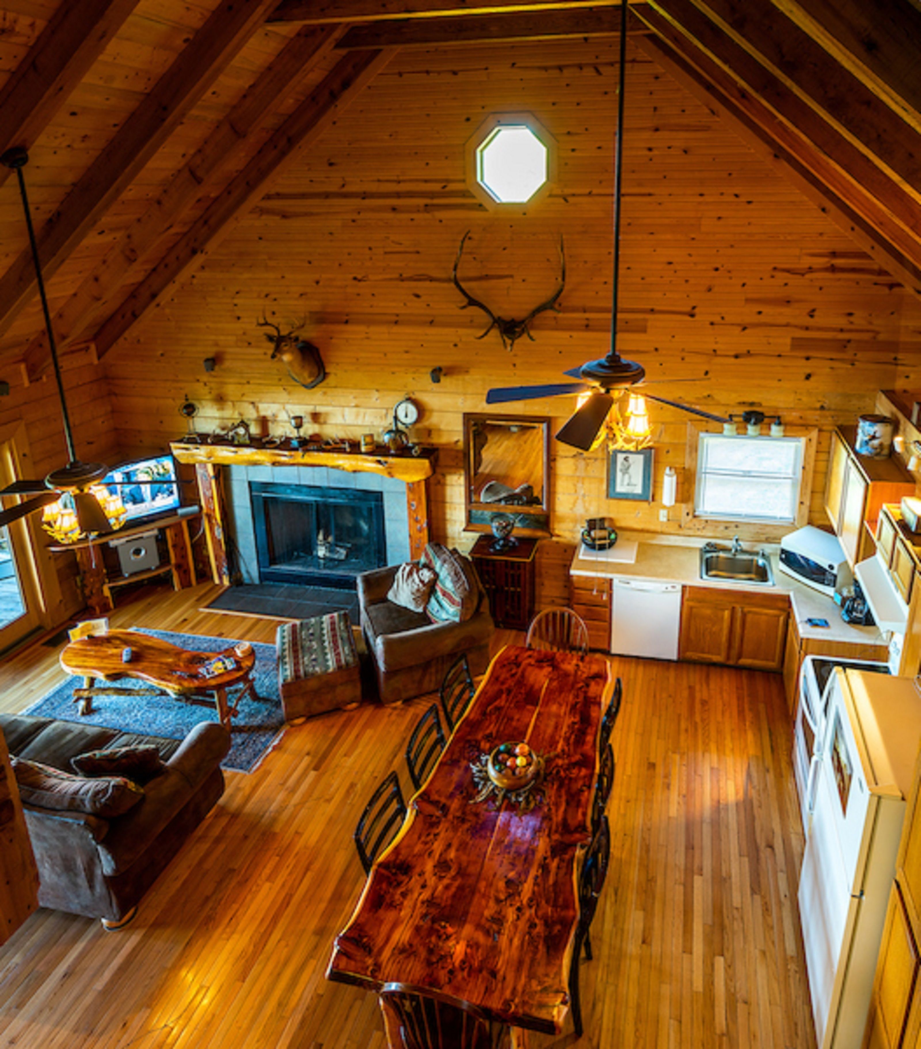 A cozy cabin interior with a wooden ceiling, a fireplace, a dining table, and a kitchen area.