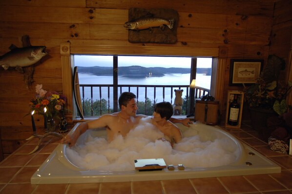 A wooden cabin with a large bathtub filled with bubbles, a wooden fish mounted on the wall, and a view of a lake through a window.