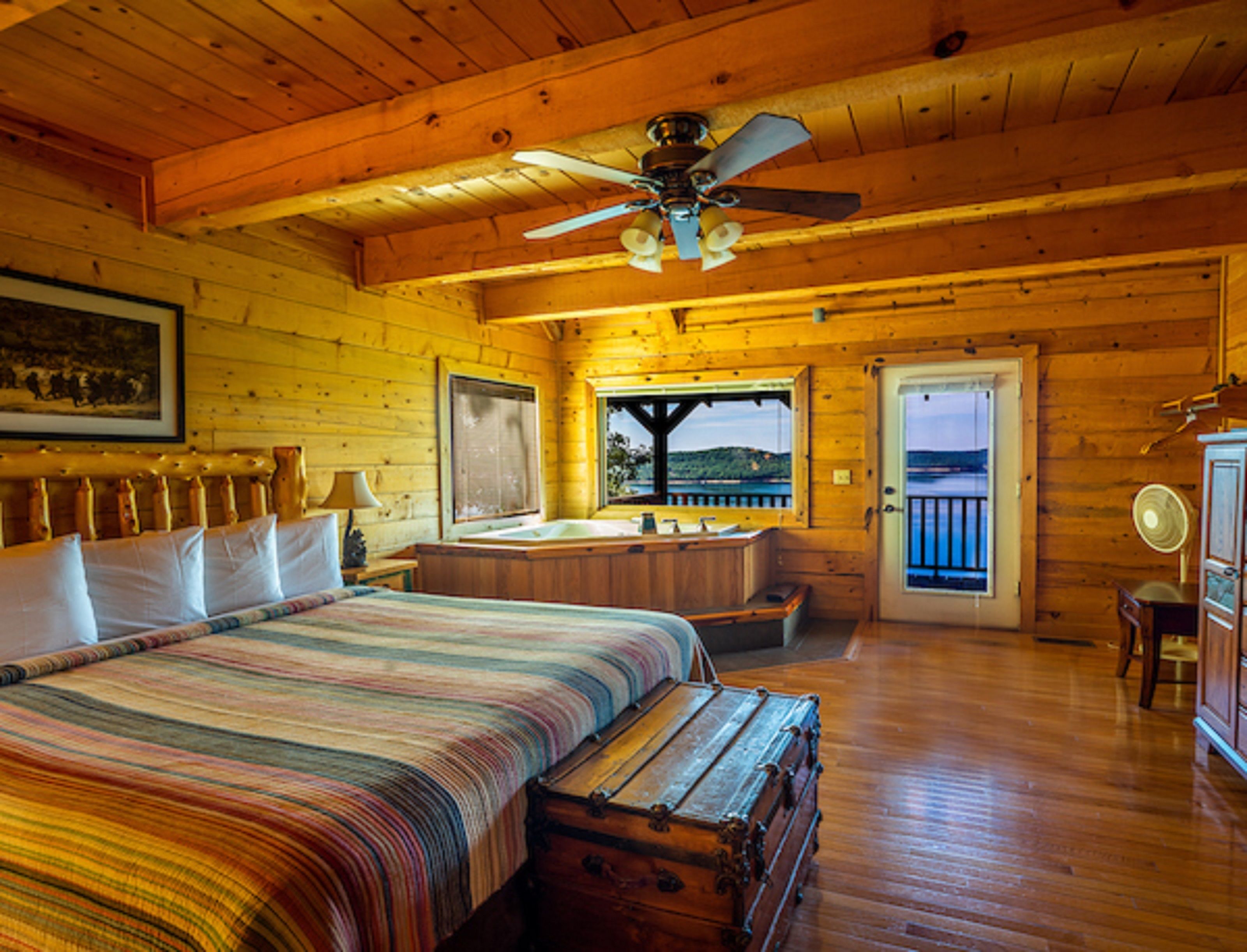 Un dormitorio de cabaña rústica con una cama grande, un ventilador de techo, vistas a través de una puerta y suelo de madera.