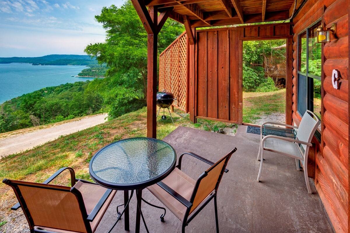 A wooden cabin with a patio featuring a glass table and chairs, overlooking a body of water and greenery.