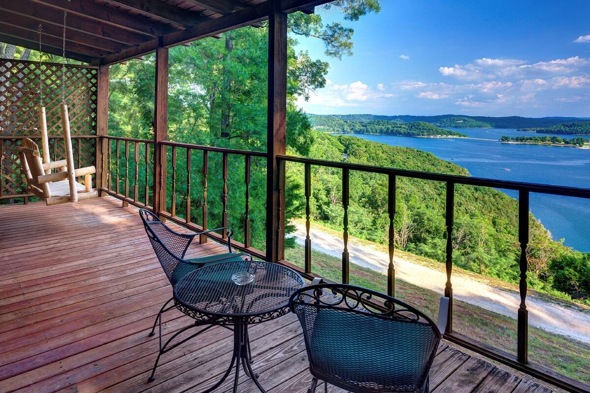 A wooden deck with two metal chairs and a table, overlooking a lake and forested hills.