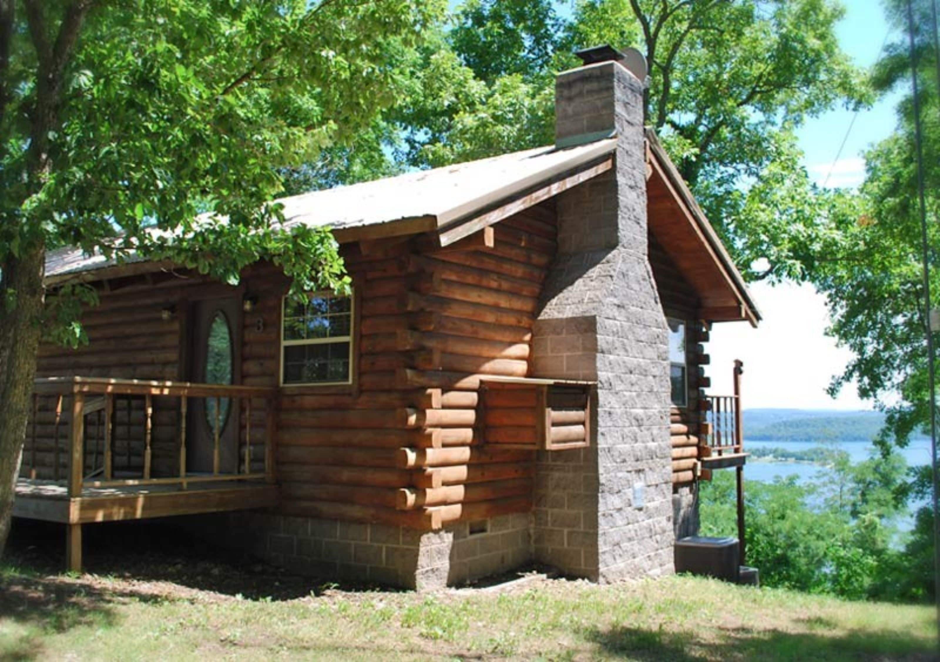 Una cabaña de troncos con chimenea de piedra, una terraza y vistas a un lago.