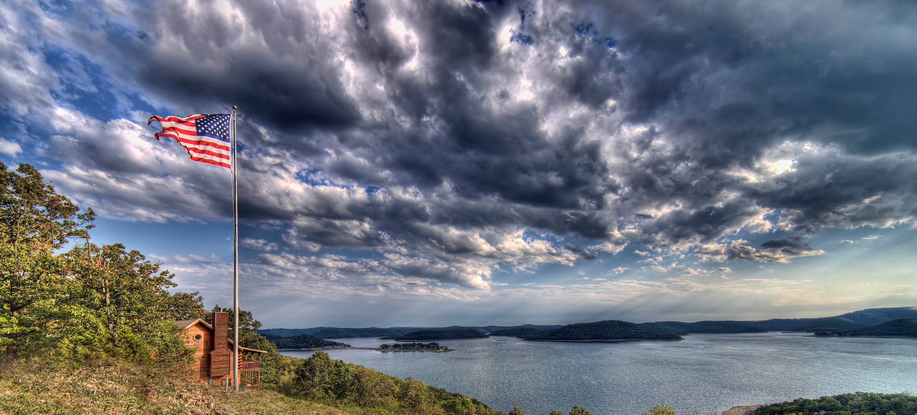 A flagpole with an American flag, a wooden cabin, and a lake with distant islands under a cloudy sky.