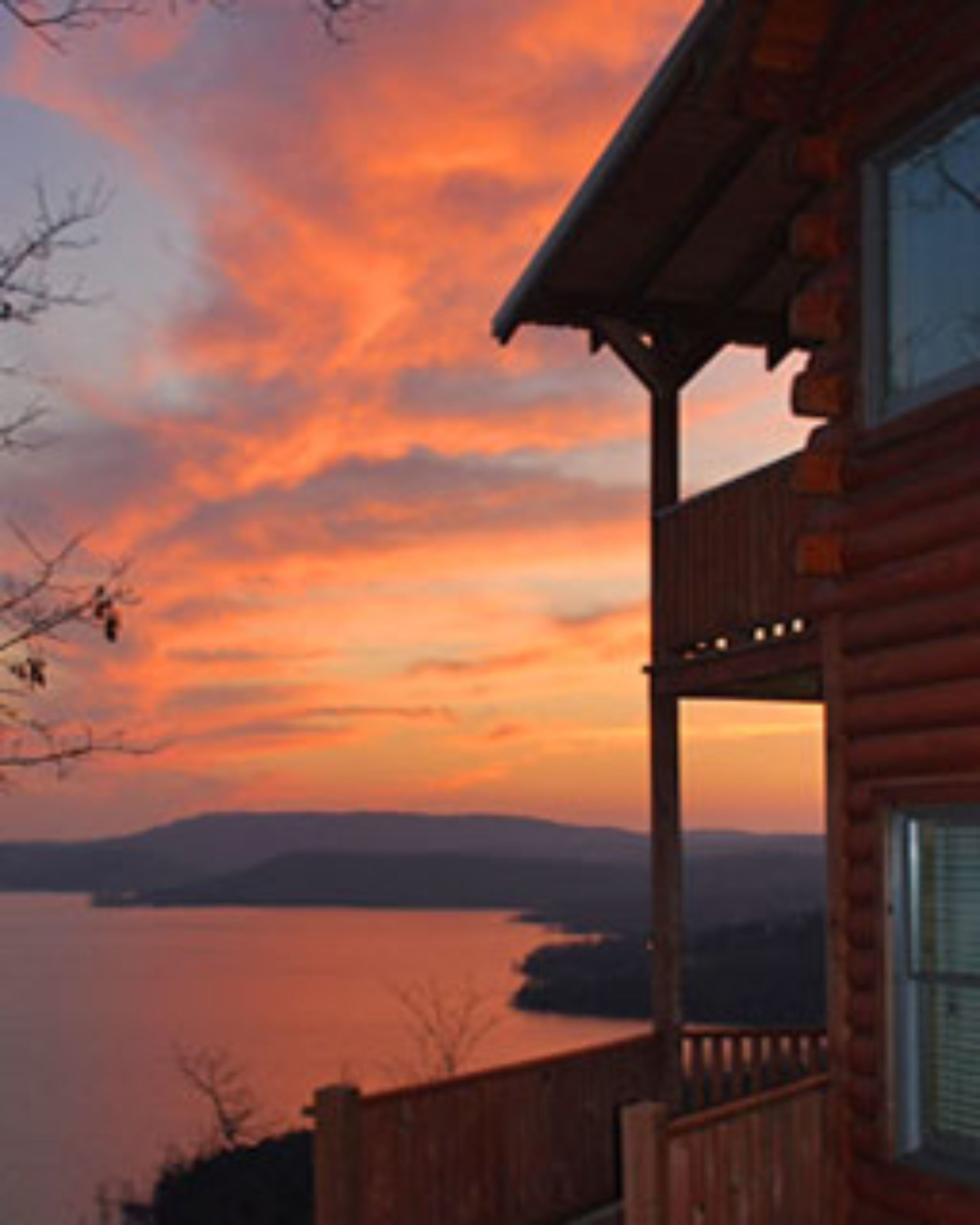 A wooden cabin with a balcony overlooking a body of water and distant hills during sunset.