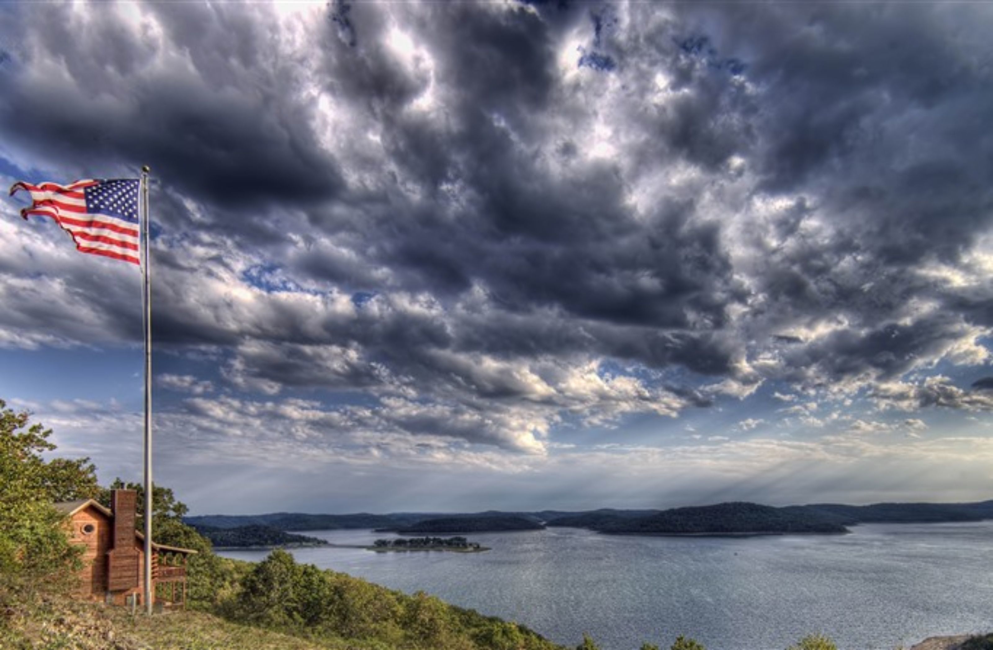 A view of a lake with a wooden cabin and an American flag, surrounded by cloudy skies.