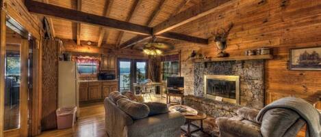 A rustic cabin interior with wooden beams, a stone fireplace, a sofa, and a television.