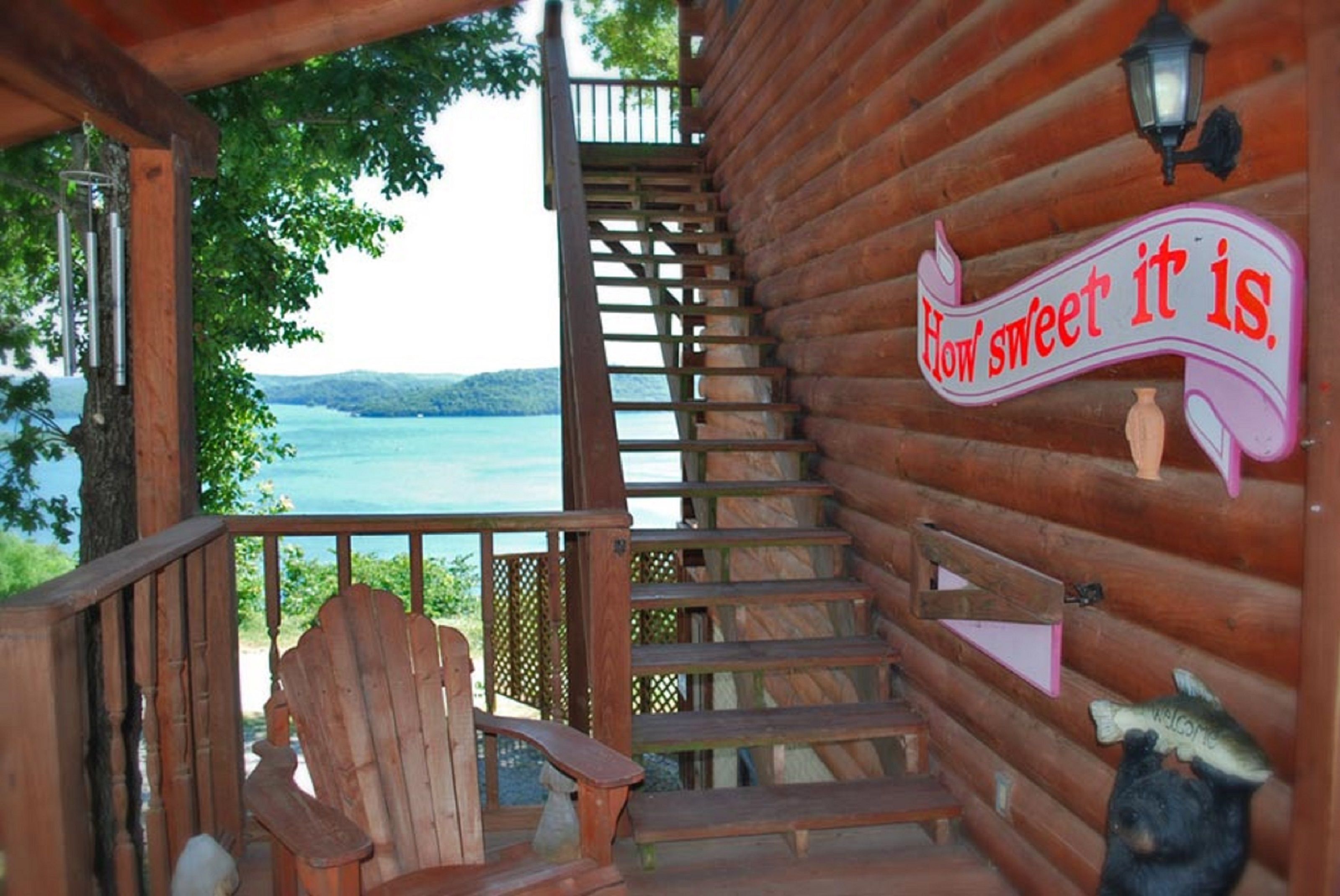 A wooden staircase leading to a deck with a view of a lake, a wooden chair, and a sign that says "How sweet it is."